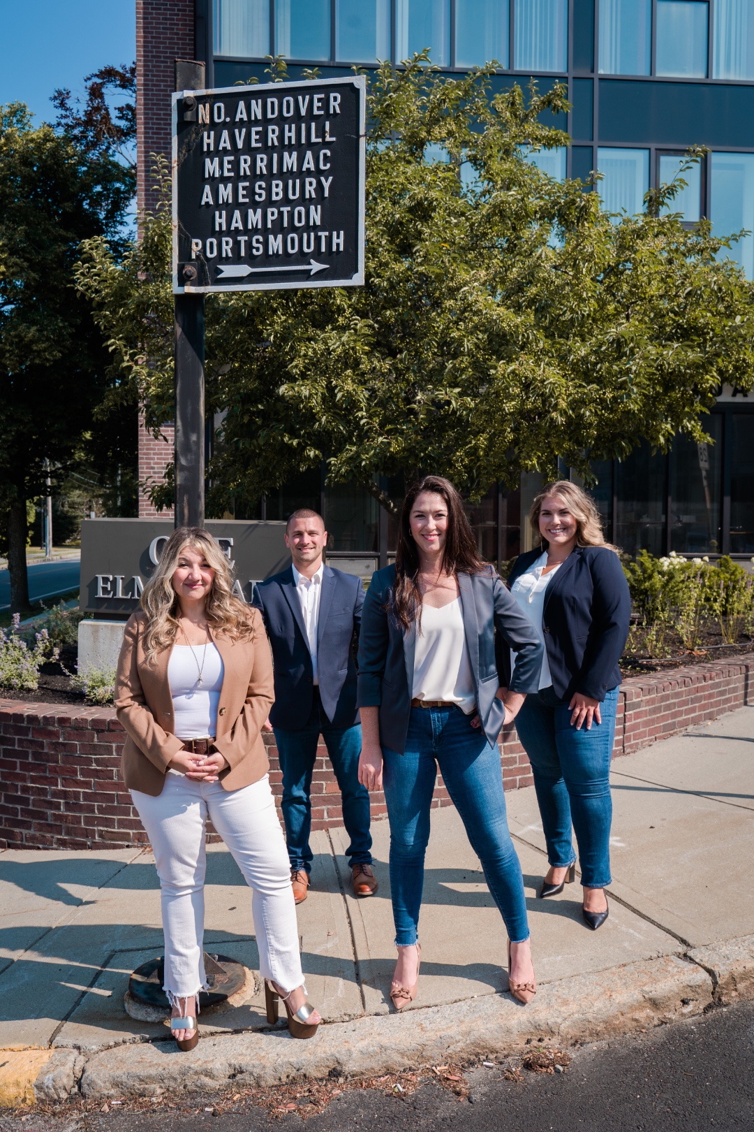 The Lisa Sevajian Group team standing under the No. Andover / Haverhill / Merrimac / Amesbury / Hampton / Portsmouth directional sign at One Elm in downtown Andover, MA
