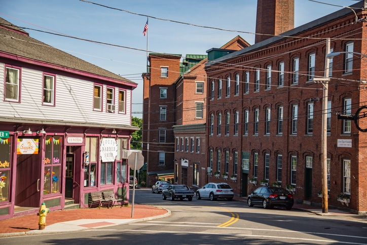 Brick mill buildings and historic storefronts on a sunny New England street.